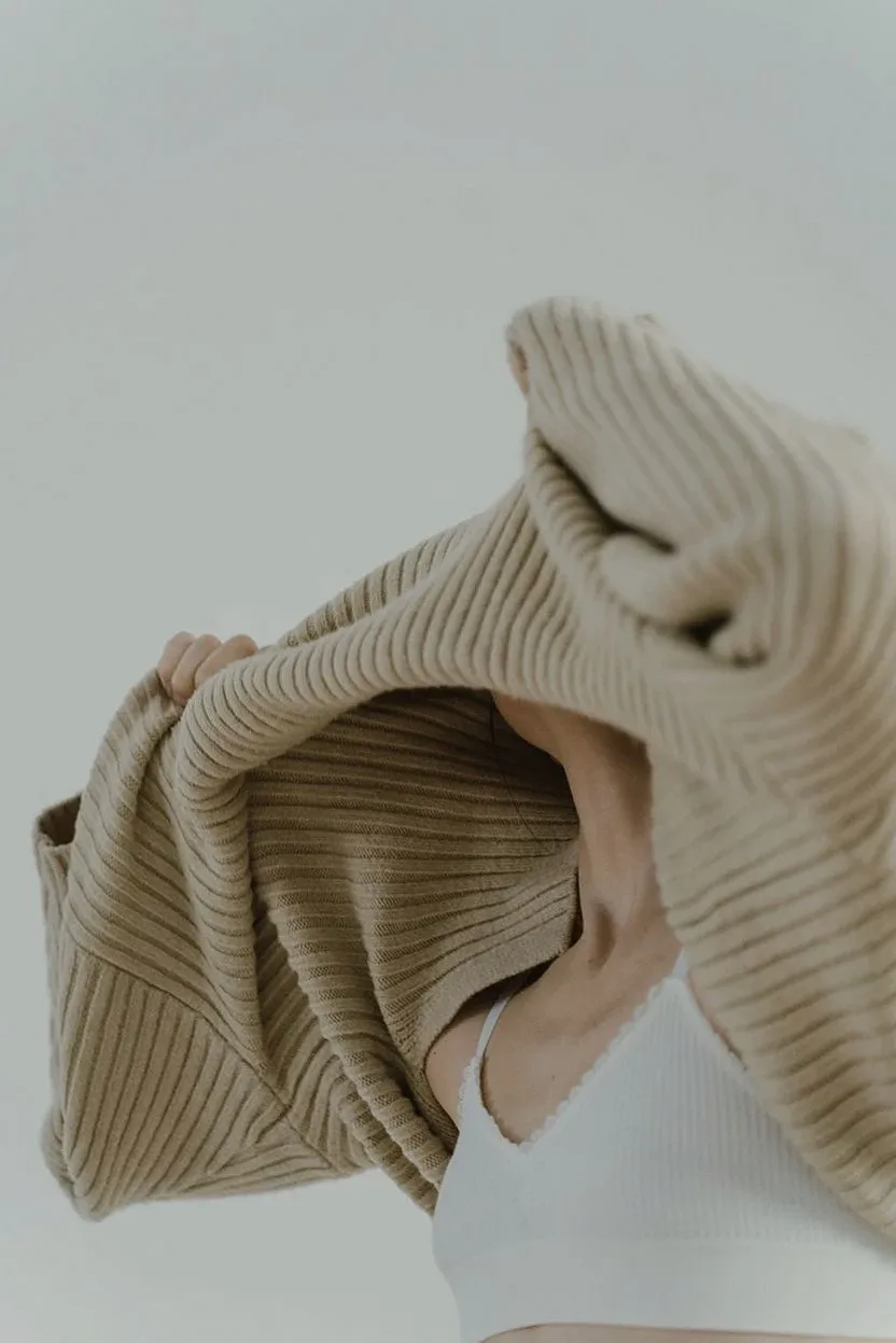 Woman pulling a beige knit jumper over a white bralette
